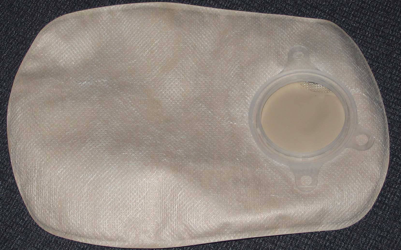 A beige ostomy pouch with a clear flange ring lying on a dark fabric surface.