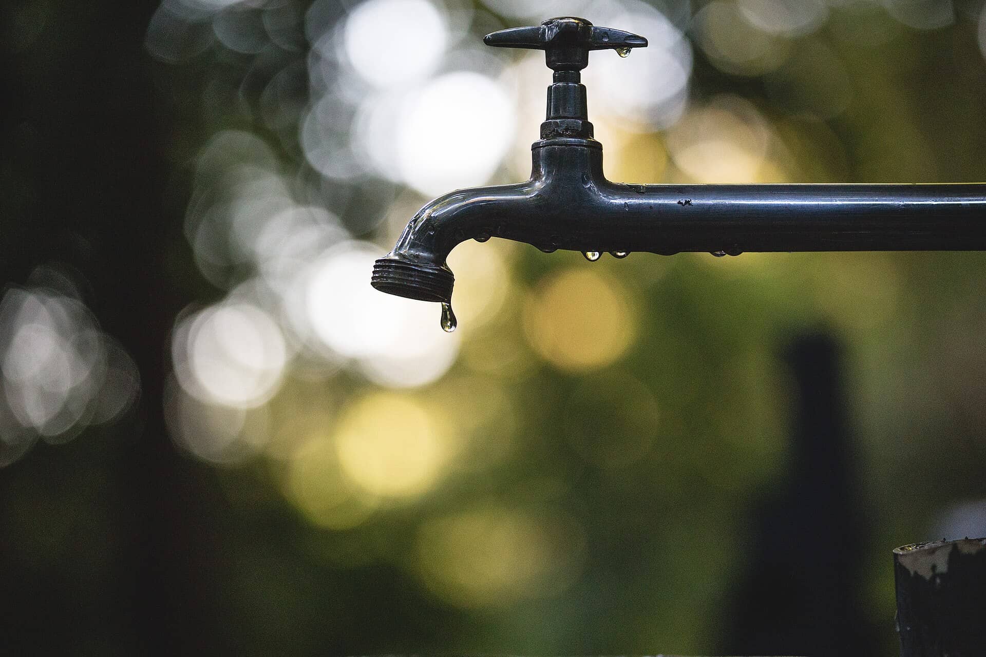 A dark metal outdoor tap with water drops, one falling from the spout.