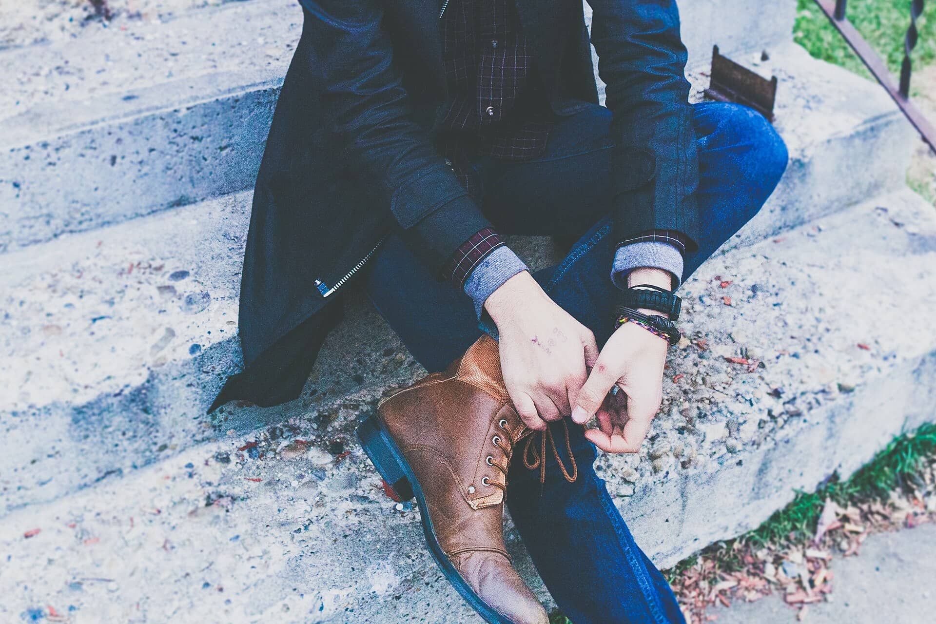 A seated person ties the laces of a brown leather boot on concrete steps; no face is visible.