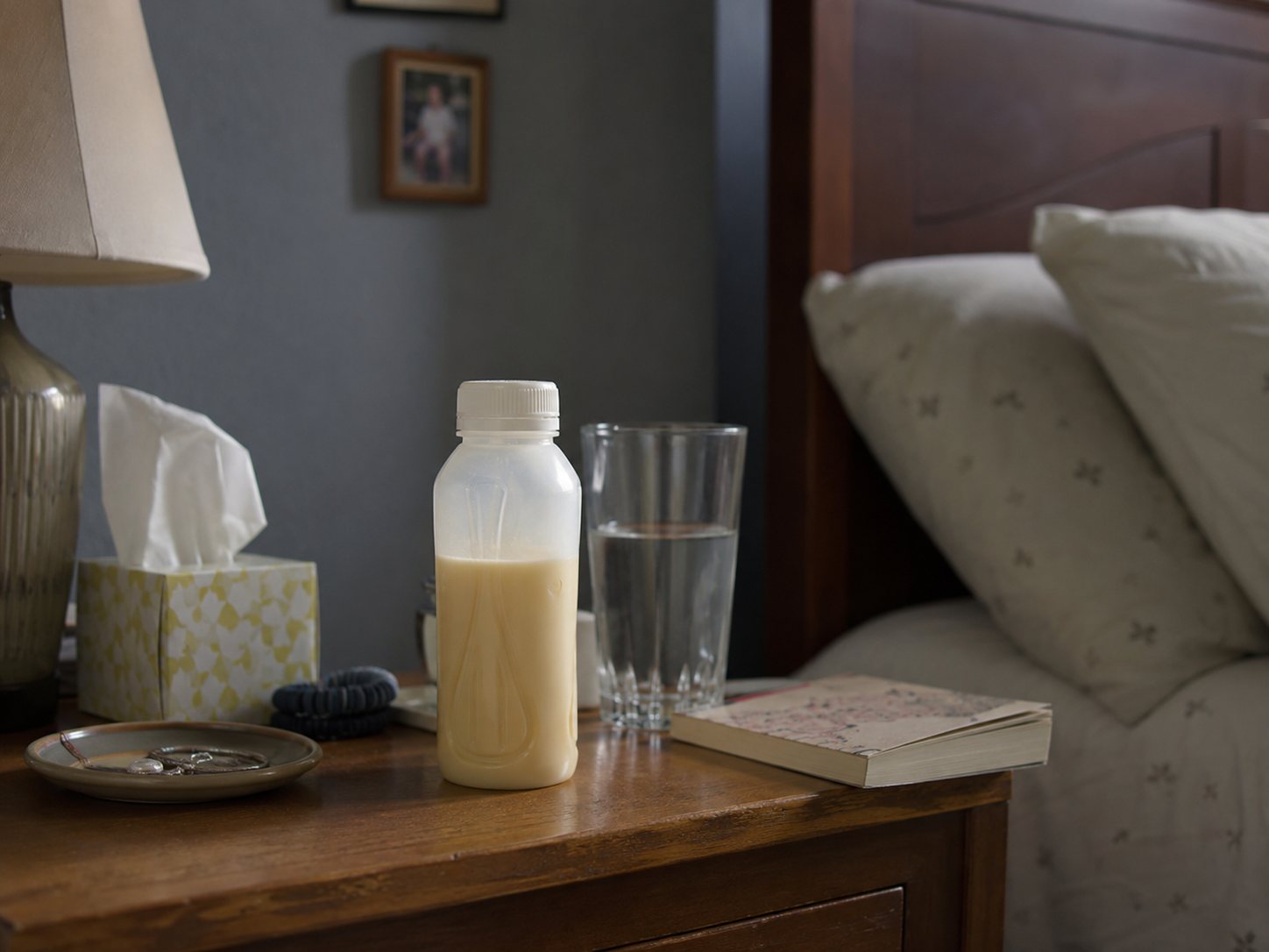A bottled nutrition drink, a glass of water, a box of tissues and a book on a wooden nightstand beside a bed