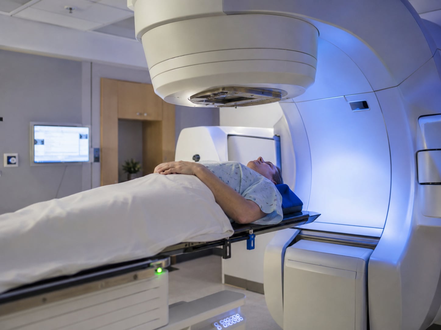A patient lies alone on the couch of a linear accelerator with the treatment head positioned above them in a blue-lit treatment room.